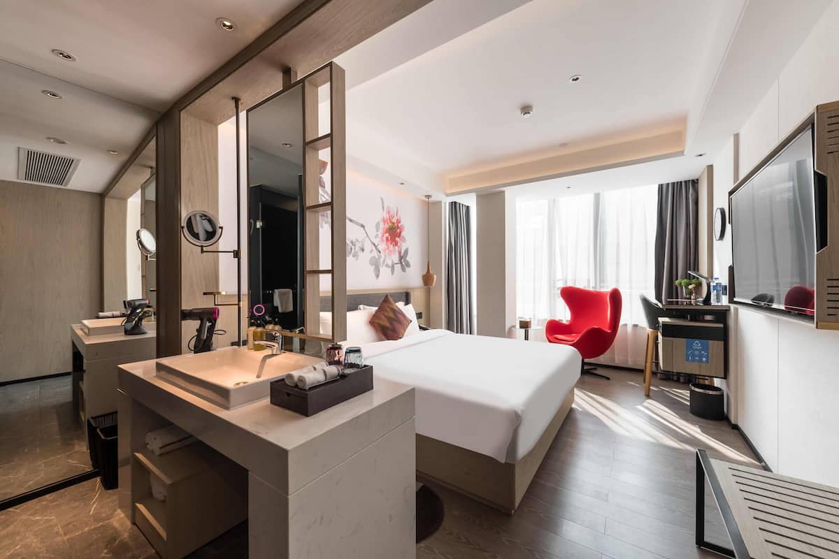 A hotel room with a large bed, a desk with a computer, a red chair, and a bathroom with a sink and mirror.