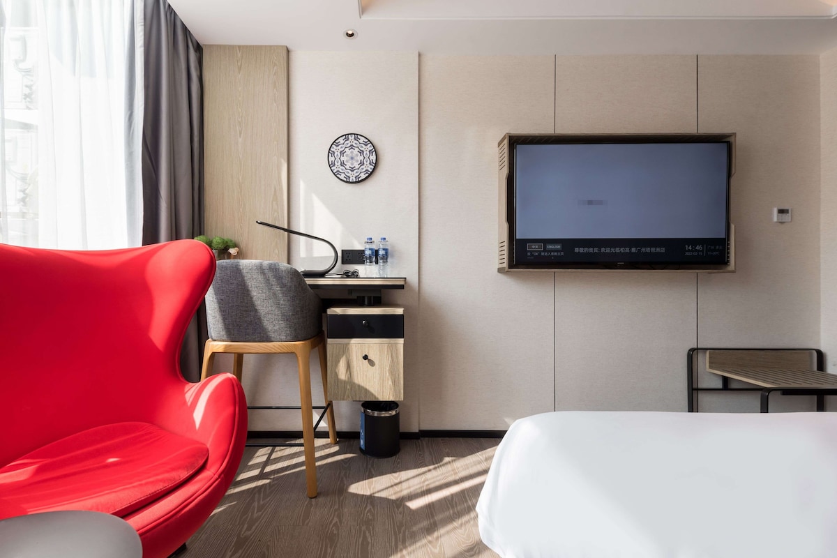 A hotel room with a bed, a red chair, a desk with a TV, and a small trash bin.