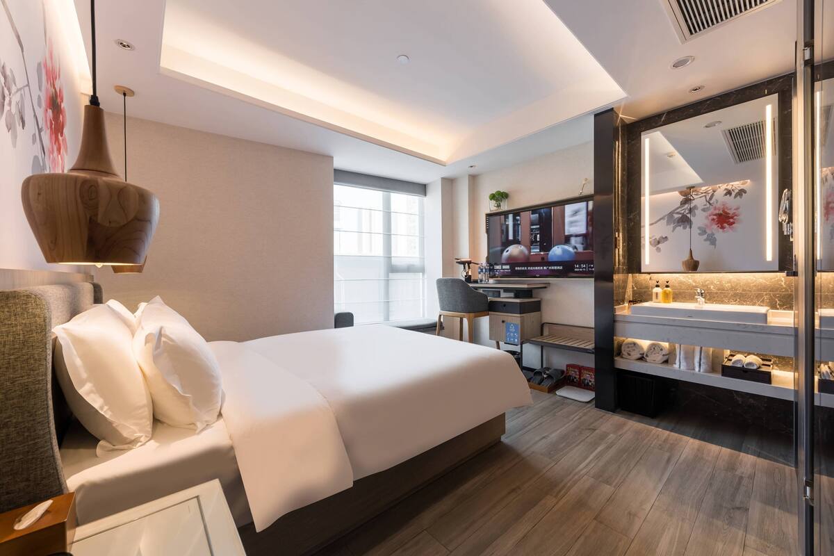 A modern hotel room with a large bed, a desk, and a bathroom with a mirror and sink.