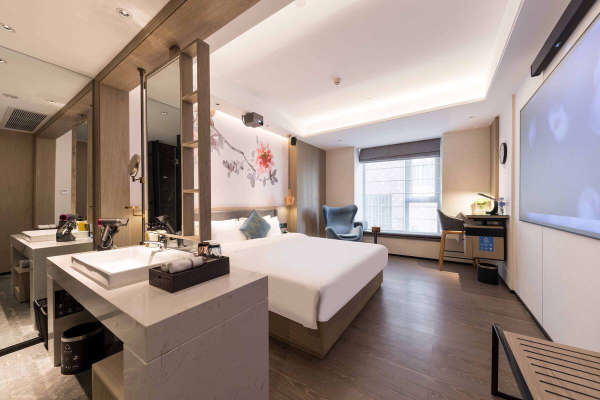 A modern hotel room with a large bed, a desk, a chair, a large mirror, and a bathroom with a sink and vanity.