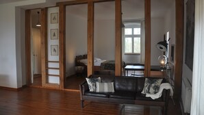 A room with a wooden floor, a black leather sofa with decorative pillows, a desk with a lamp, and a bed with a window.