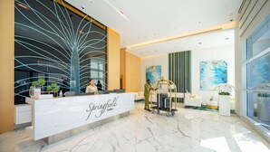 Una moderna zona de recepción de hotel con suelo de mármol, una mesa de recepción que dice "Springfield Suites" y un carrito de equipaje.