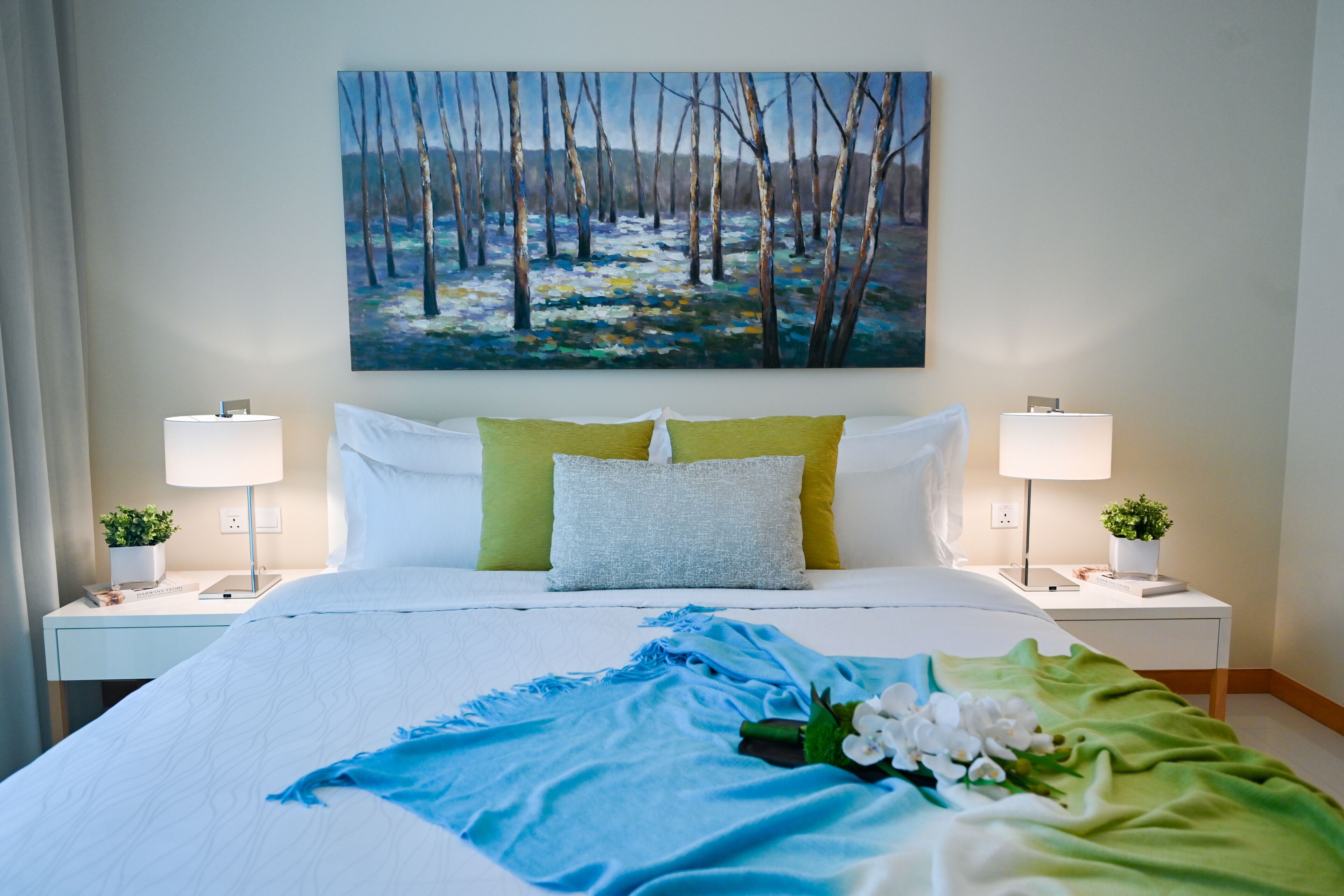 A neatly made bed with a blue and green blanket, white pillows, and a painting of a forest on the wall.