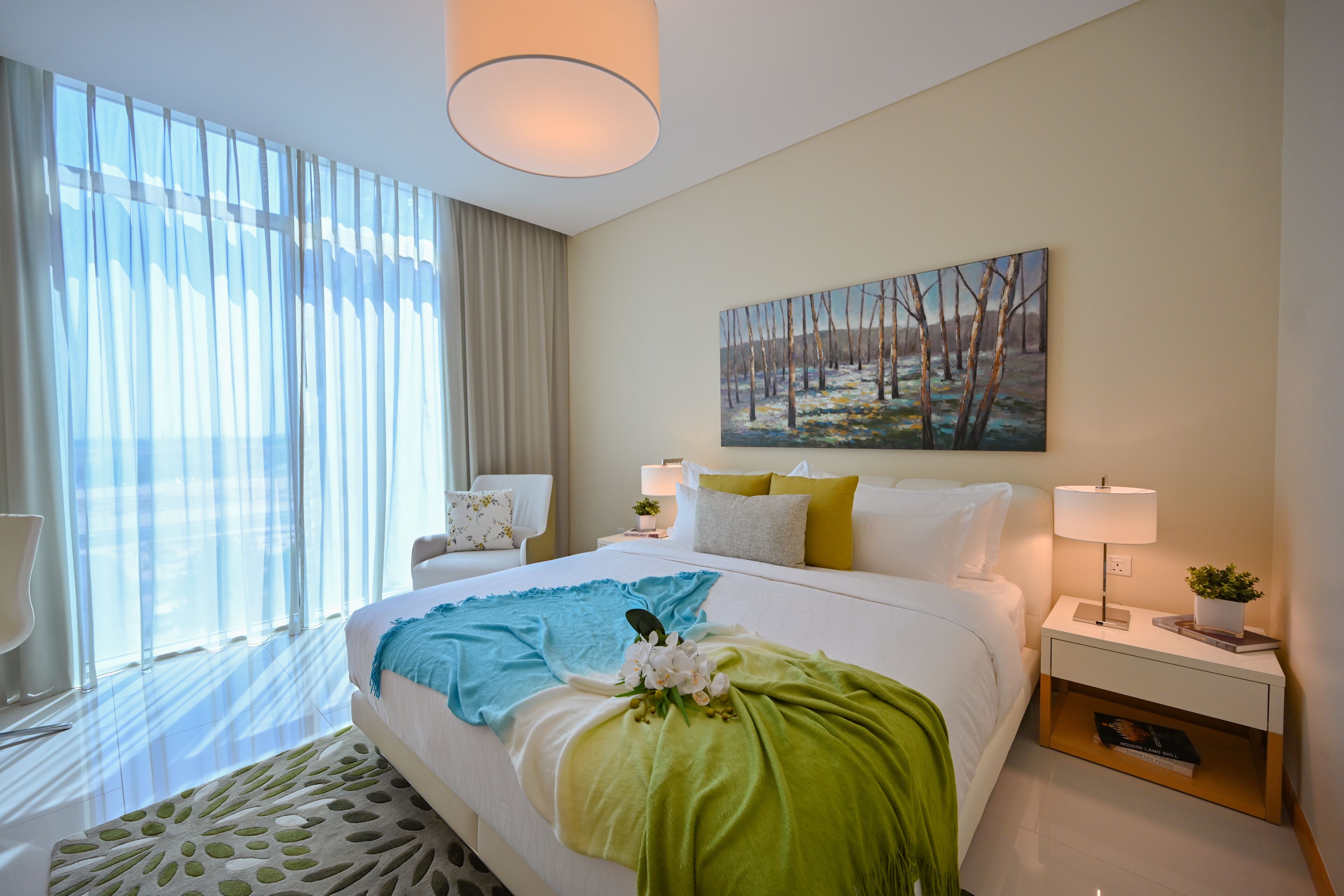 A hotel room with a large bed, a painting on the wall, a nightstand with a lamp, and a window with curtains.