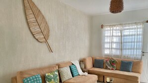 Una sala de estar con un sofá seccional color beige, un adorno de hojas de madera y una ventana con cortinas traslúcidas.