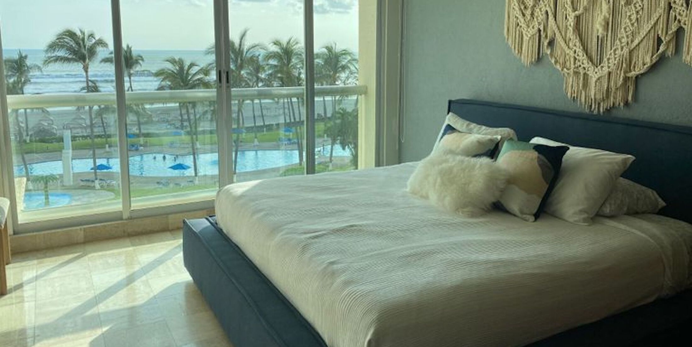 Quarto com cama grande, vista para piscina e palmeiras, e um painel de macramé na parede.