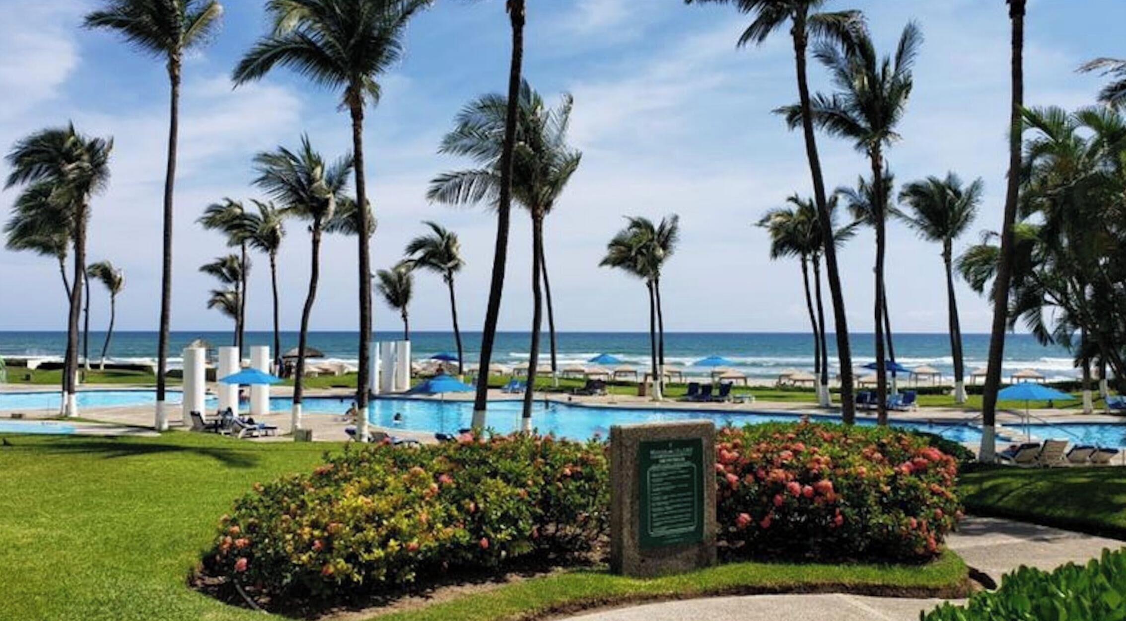 Área de piscina em resort, com palmeiras, céu claro e vista para o oceano.
