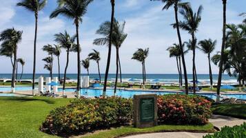 Área de piscina em resort, com palmeiras, céu claro e vista para o oceano.