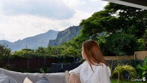 Une personne est assise sur un canapé gris, avec vue sur un paysage montagneux et une végétation luxuriante.