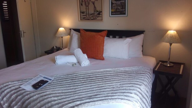 A neatly made bed with a striped blanket, two pillows, and a folded towel. There are two bedside lamps, a framed picture on the wall, and a door in the background. - Soeteweide North Bb - Close to Fancourt Golf Estate (George)