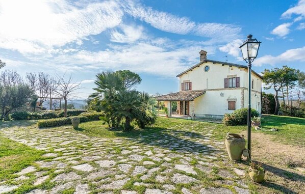 A white building with a tiled roof, surrounded by a stone-paved courtyard and lush greenery.