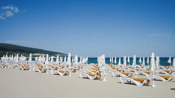 A beach with rows of sun loungers and umbrellas, a clear sky, and a distant view of a town. - Seaside Apartment With 2 Bedrooms for up to 5 Guests (Nessebar)