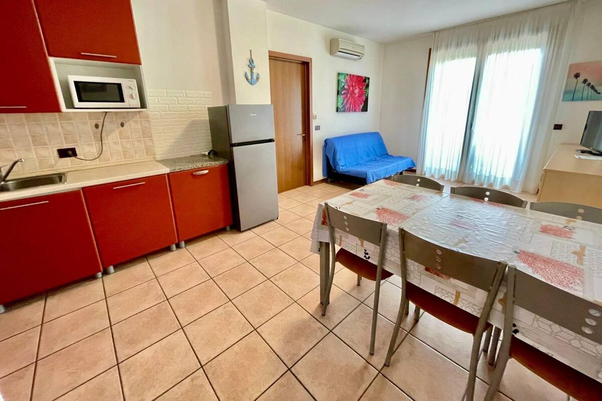 A compact kitchen with red cabinets, a microwave, and a refrigerator. There is a dining table with chairs and a blue sofa in the adjacent living area.