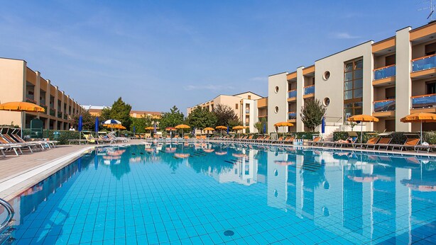 Une piscine dont le fond est recouvert de carrelage, entourée de chaises longues et de parasols, et située à côté d’immeubles à plusieurs étages dotés de balcons.