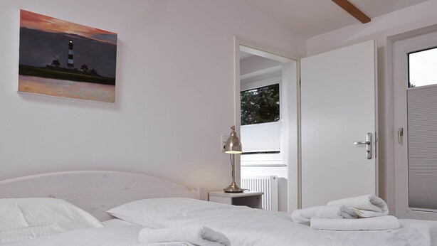 A hotel room with two beds, a painting of a lighthouse, a lamp, and a view of the outside through an open door. - Birklund - Holiday home for two (Sankt Peter-Ording)