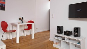 A modern living room with a white TV, black speakers, a red chair, a white table, and a wooden floor. - Birklund - Holiday home for two (Sankt Peter-Ording)