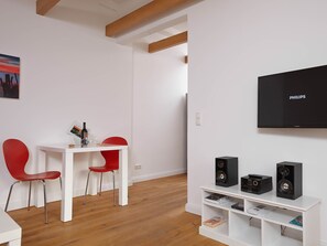 A modern living room with a white TV, black speakers, a red chair, a white table, and a wooden floor.