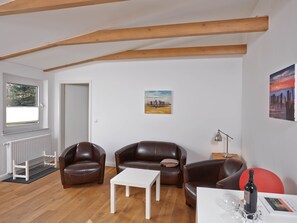 A living room with wooden beams, a white radiator, a window, two brown leather sofas, a white coffee table, a red chair, a floor lamp, and a wine bottle.
