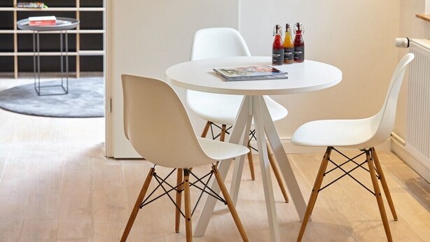 Een moderne eethoek met een ronde tafel en vier witte stoelen met houten poten. Er staan drie flessen op de tafel, en er ligt ook een tijdschrift.