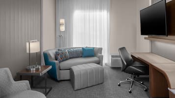 A modern hotel room with a grey sofa, a small table with a lamp, a desk with a chair, and a large flat-screen TV mounted on the wall.