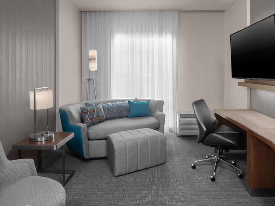 A modern hotel room with a grey sofa, a small table with a lamp, a desk with a chair, and a large flat-screen TV mounted on the wall.
