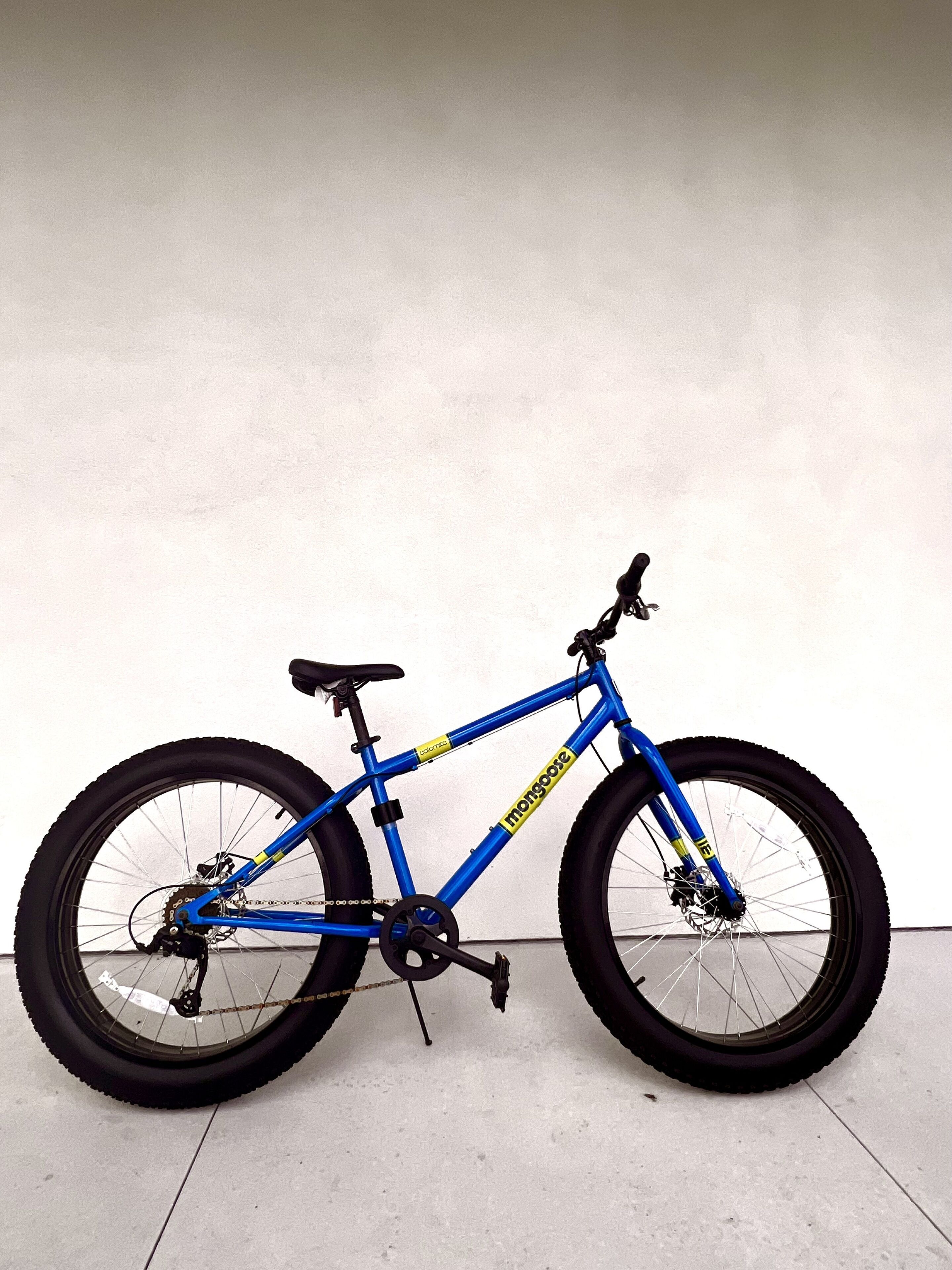 A blue bicycle with a yellow label, black seat, and black tires, leaning against a white wall.