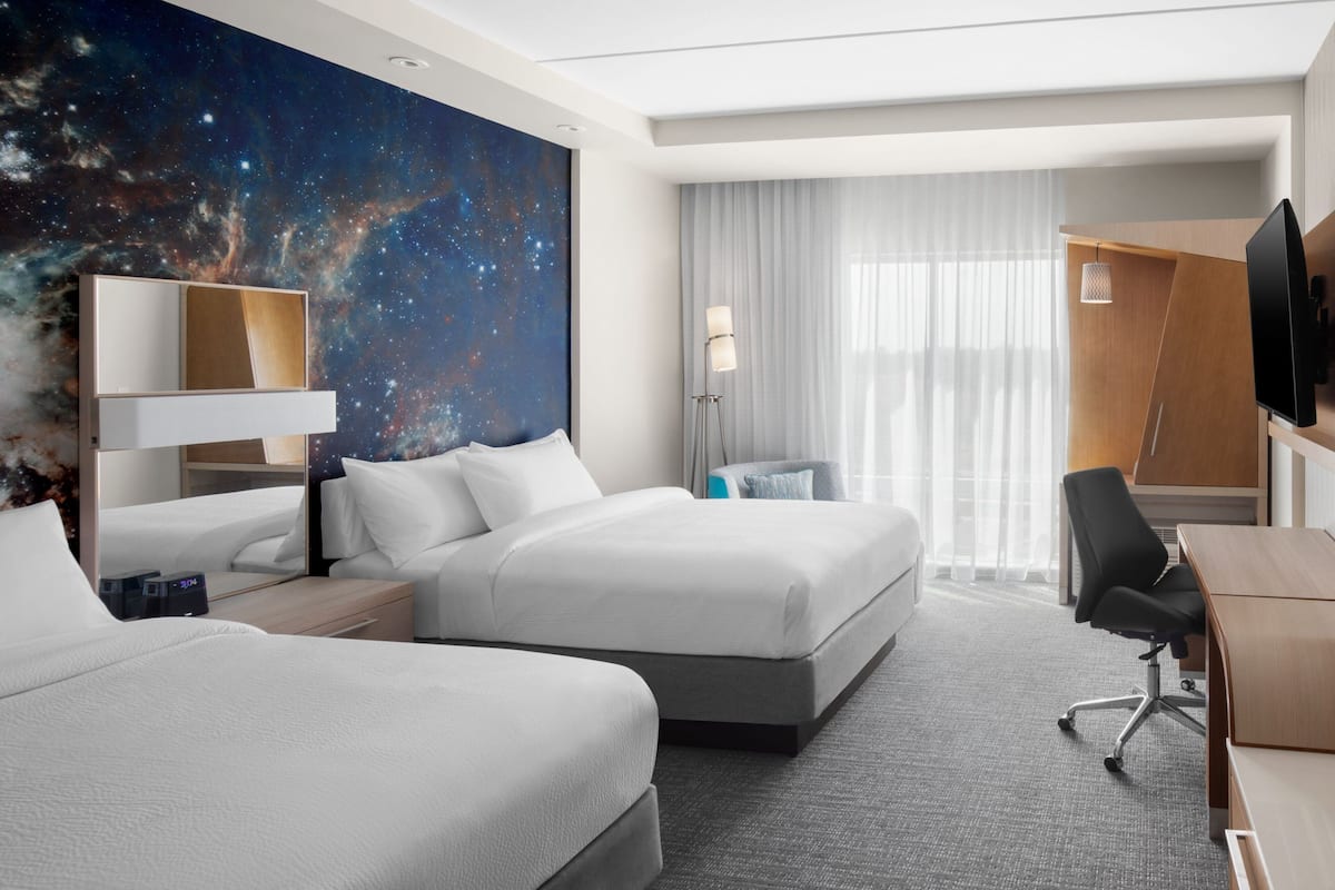 A hotel room with two beds, a desk, a chair, a TV, a large window with curtains, and a galaxy-themed wall mural.
