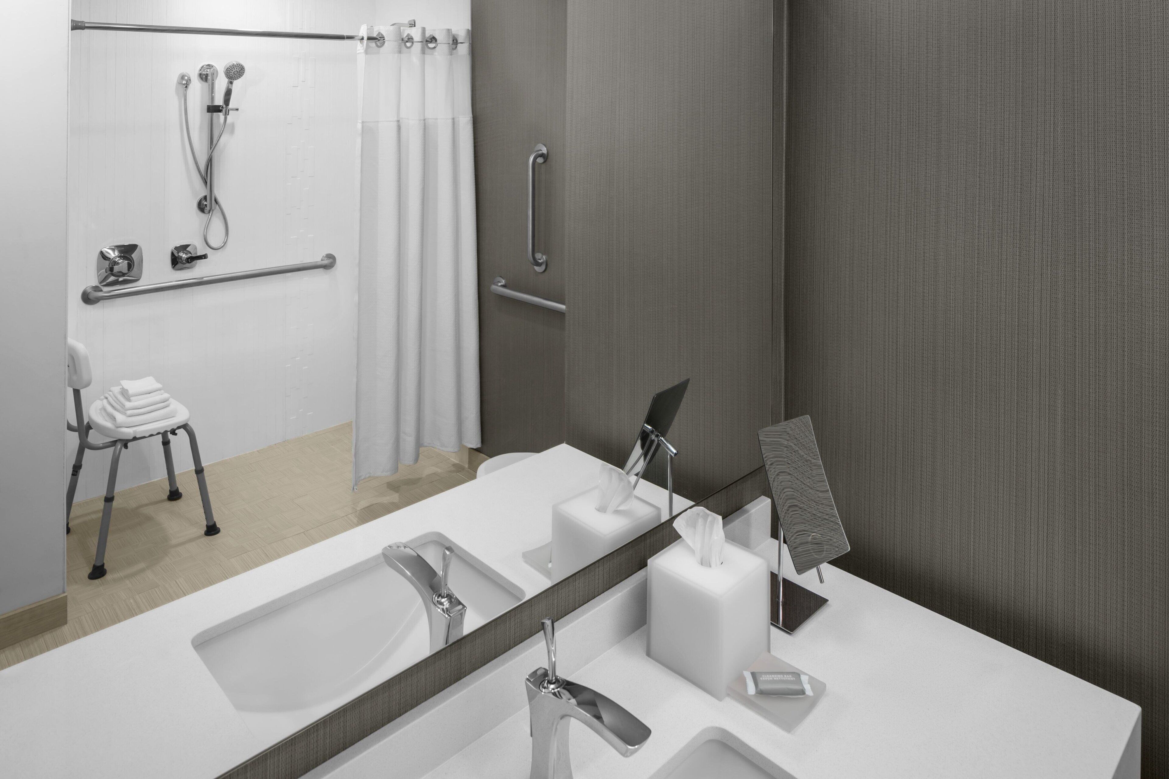 A modern bathroom with a large mirror, two sinks, a shower with a handheld showerhead, a towel rack, and a grab bar.