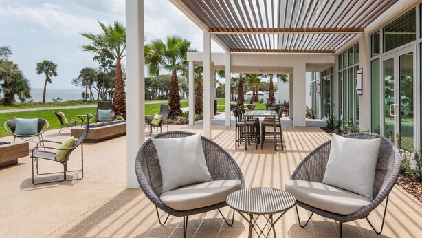 An outdoor patio with wicker chairs and a table, overlooking a landscaped area with palm trees and a view of the sea. - Courtyard by Marriott Titusville Kennedy Space Center (Titusville)