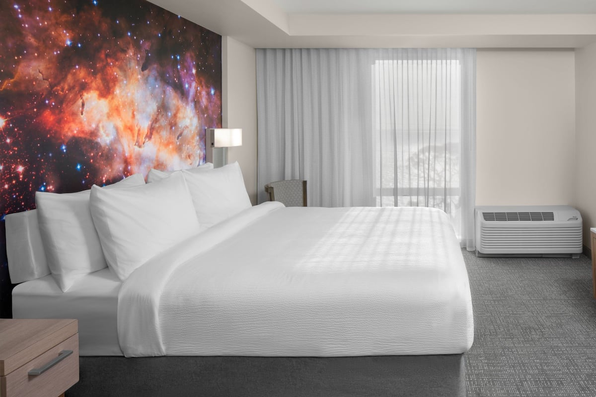 A hotel room with a large bed, a nightstand, a desk, a chair, a window with curtains, and a wall with a space-themed mural.