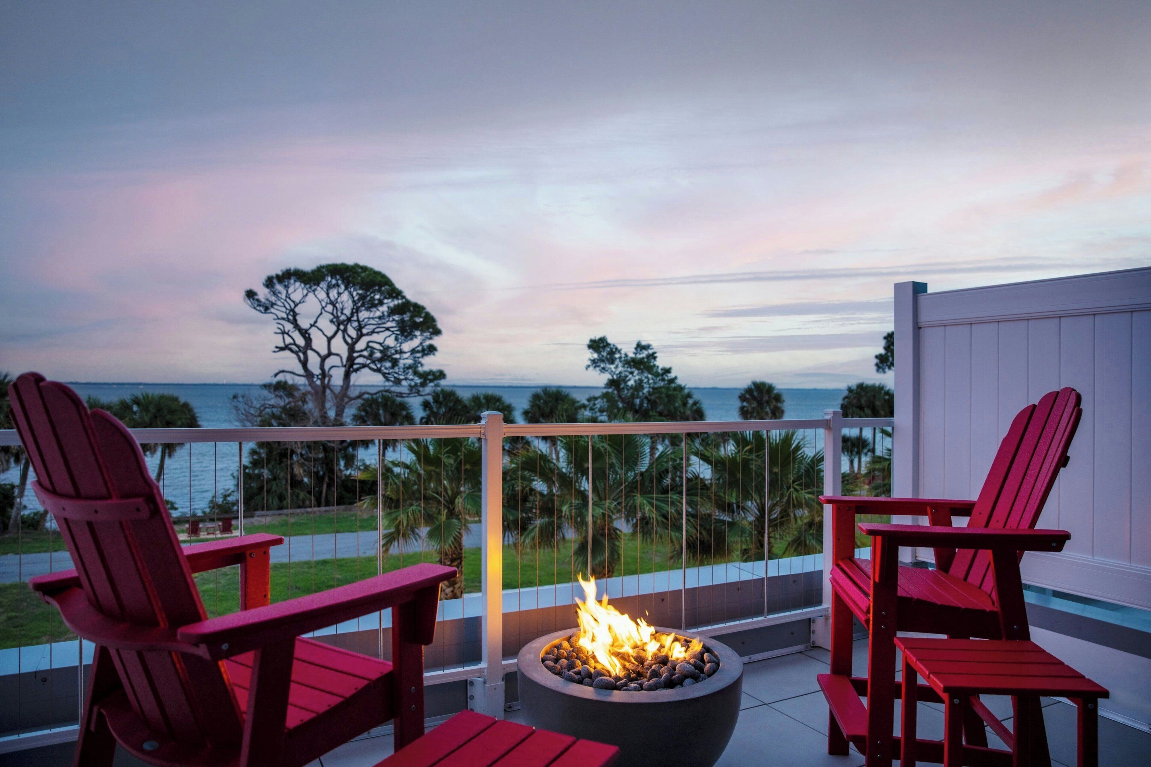 A balcony with a fire pit, two red chairs, and a view of the ocean at dusk.
