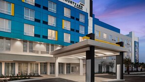 A modern hotel with a blue and yellow facade, a covered entrance, and a landscaped area with palm trees. - Courtyard by Marriott Titusville Kennedy Space Center (Titusville)