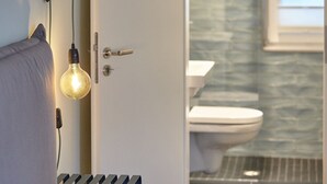 A bedroom with a bed, a bedside lamp, and a door leading to a bathroom with a toilet and a shower. - Strandpirat - Holiday apartment for two (Sankt Peter-Ording)