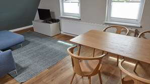 A room with a wooden dining table, four chairs, a blue sofa, a grey rug, a white cabinet with a television, and two windows with blinds.