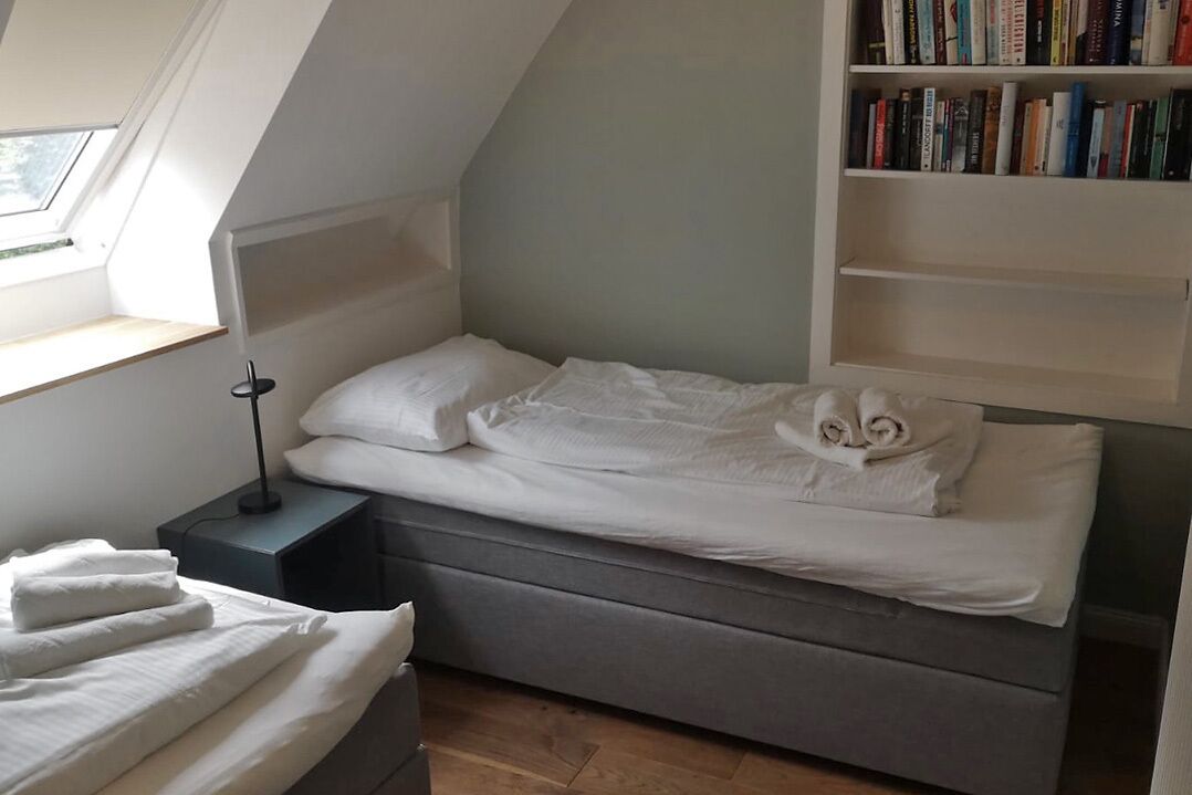 A small, neatly made bed with white linens and a rolled towel, a bedside table with a lamp, and a bookshelf filled with books.