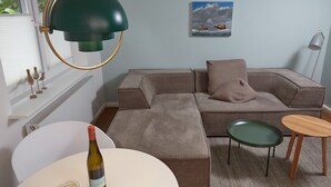 Un salon moderne avec un canapé d’angle, une table ronde sur laquelle se trouve une bouteille, un lampadaire vert et un tableau accroché au mur.