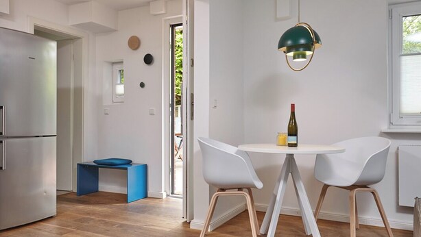 Une cuisine moderne avec un réfrigérateur en acier inoxydable, un tabouret bleu, des chaises blanches et une table sur laquelle se trouve une bouteille de vin.