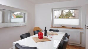 A dining area with a white table, black chairs, and a red mug. There are windows with blinds, a radiator, and a wooden shelf above the table. - Stylish vacation home Düne 10 - for up to 6 people (Sankt Peter-Ording)