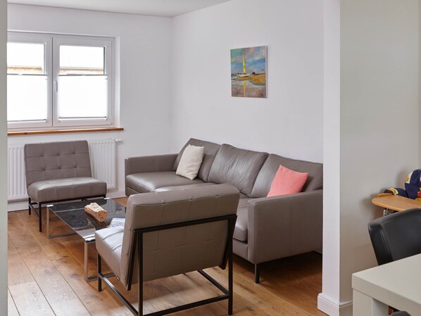 Un salon moderne avec un canapé gris, un fauteuil plus petit et une table basse. Il y a un tableau au mur et une fenêtre avec des stores.