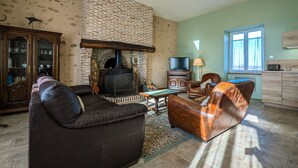 A living room with a fireplace, leather sofas, a television, and a kitchen area with a window. - Cottage in Brenne Park with garden, terrace and parking (Migné)