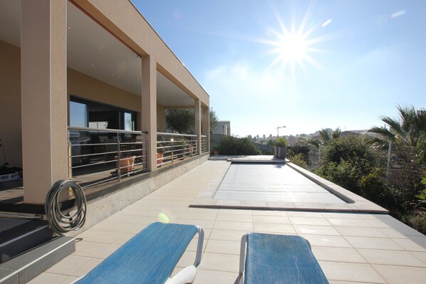 A rooftop terrace with two lounge chairs, a pool, and a building with a covered area.