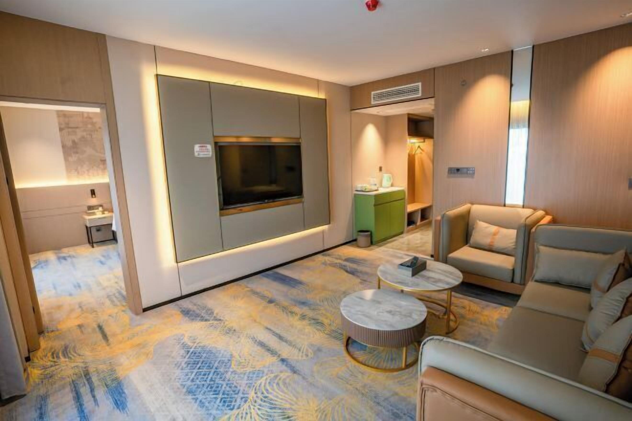 A modern hotel room with a large flat-screen TV, a sofa, a coffee table, and a patterned carpet.
