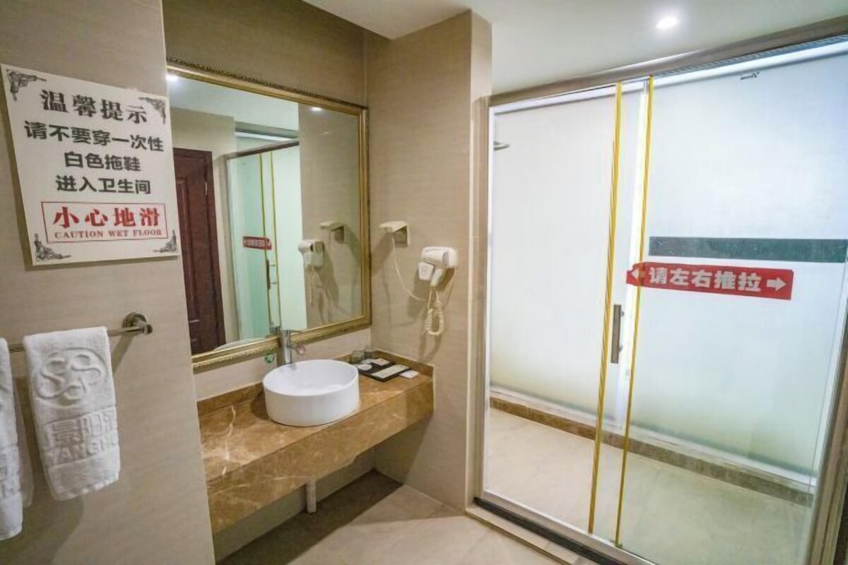 A bathroom with a white sink, a mirror, and a glass shower door. There are towels hanging on the wall and a caution sign indicating a wet floor.