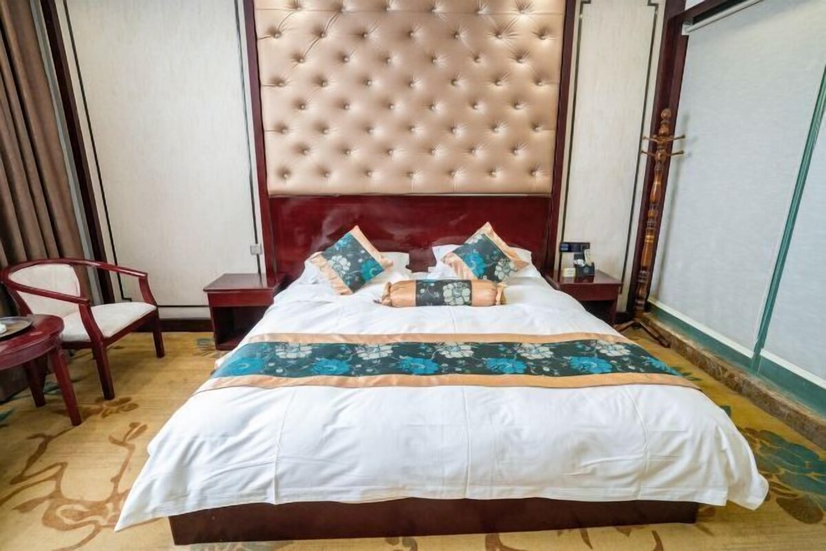 A hotel room with a tufted headboard, a bed with floral bedding, a wooden nightstand, a chair, and a small table with a bottle.