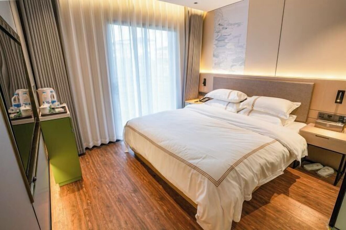 A hotel room with a large bed, a bedside table, a green cabinet, and a window with curtains.