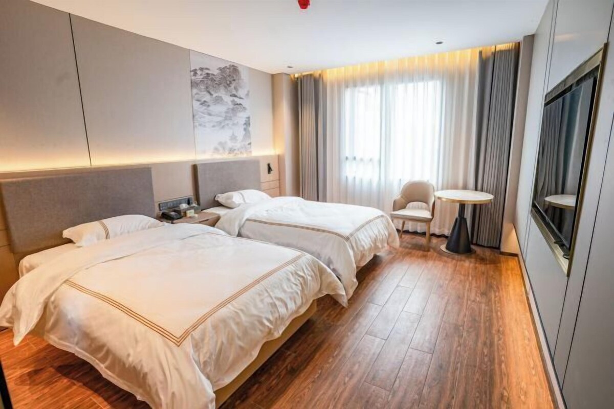 A hotel room with two beds, a small round table, a chair, a TV, and a large window with curtains.