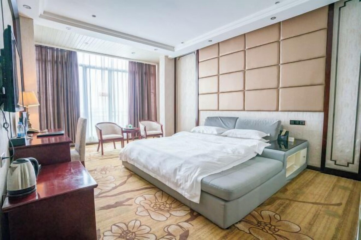 A hotel room with a large bed, a desk with a kettle, a chair, and a TV.