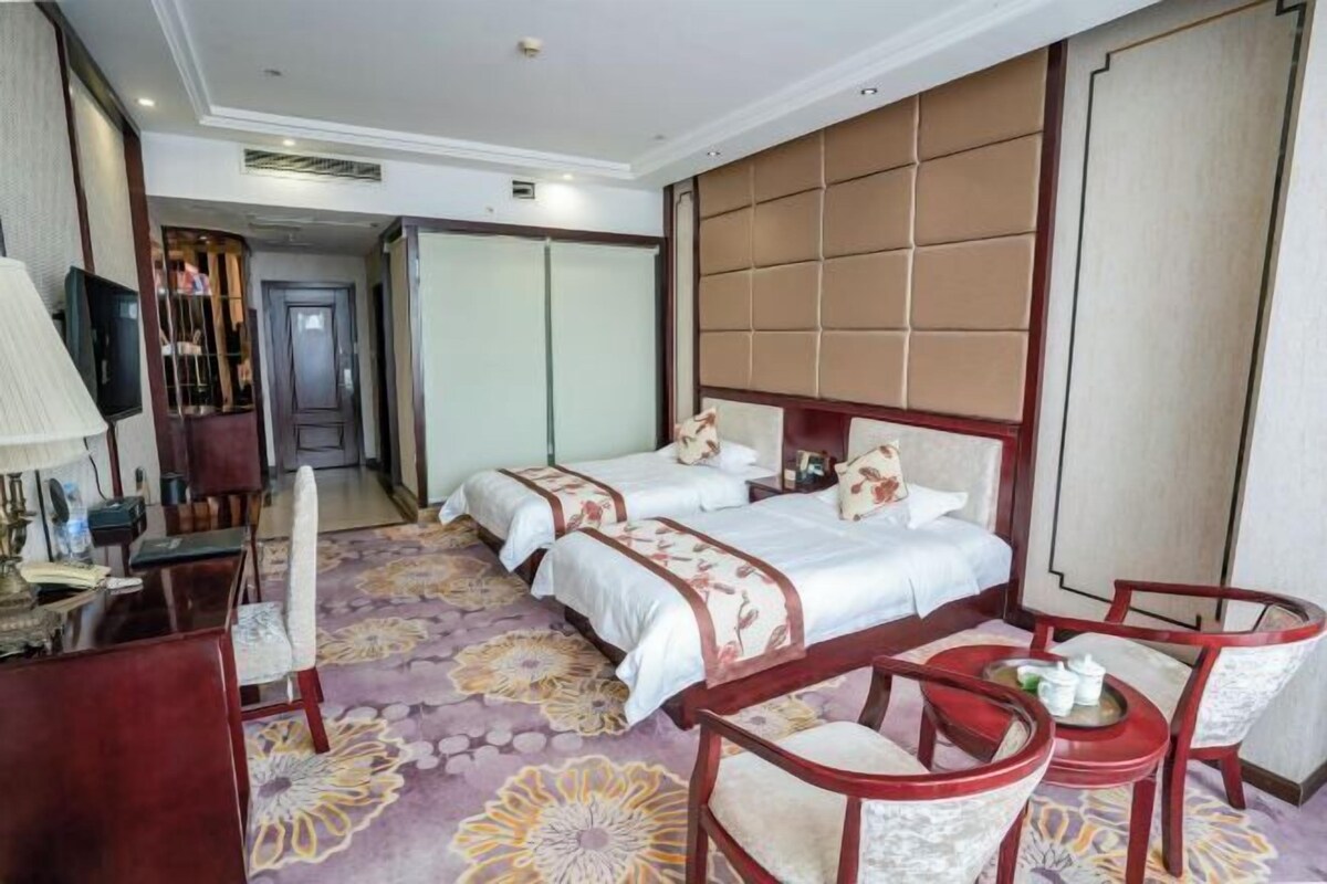 A hotel room with two beds, a desk, chairs, a television, and a decorative wall panel.