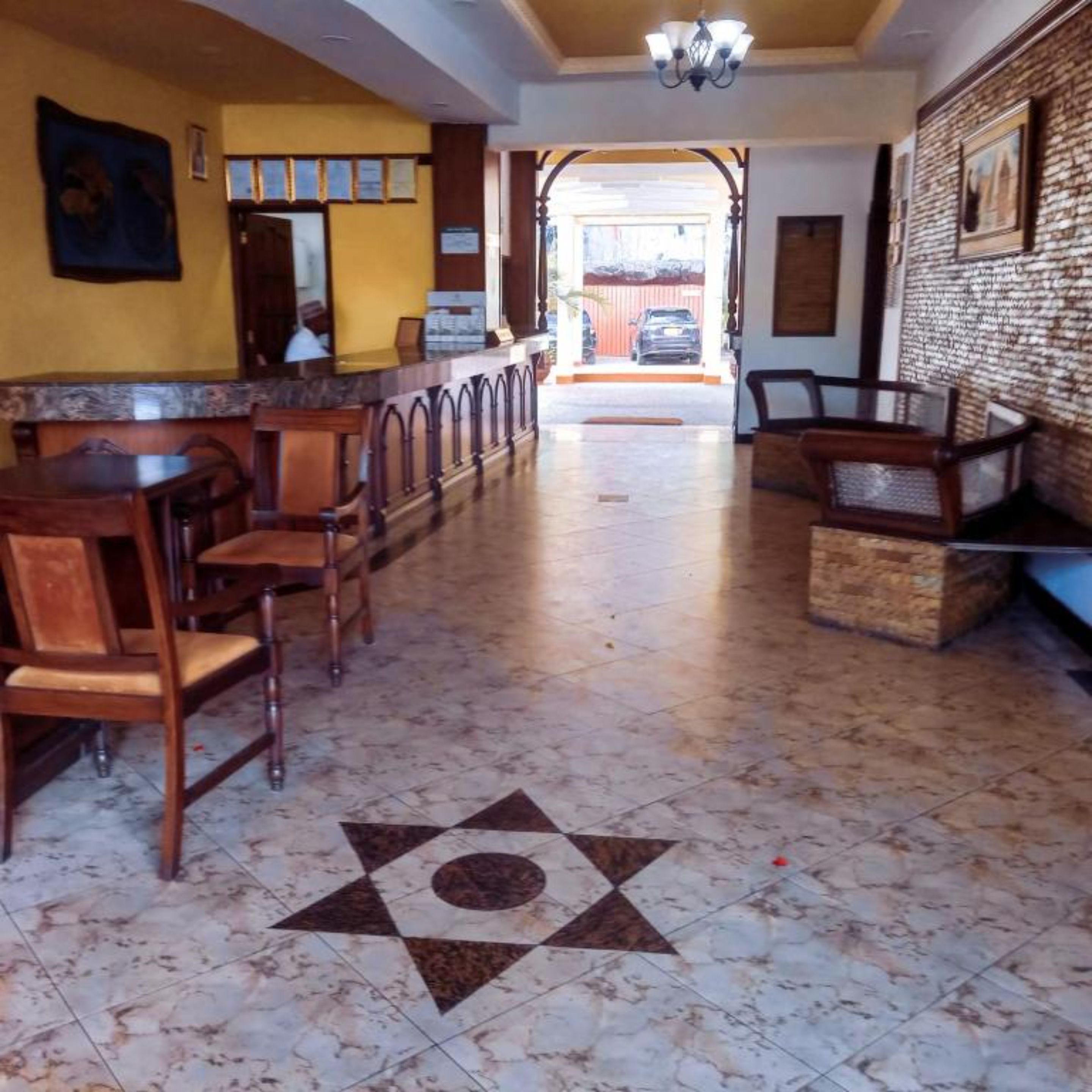A hotel lobby with a reception desk, wooden chairs, a patterned floor, and a view of the outside.