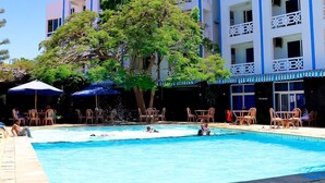 Una piscina con sillas de descanso y sombrillas, rodeada por un edificio con balcones y un árbol grande.